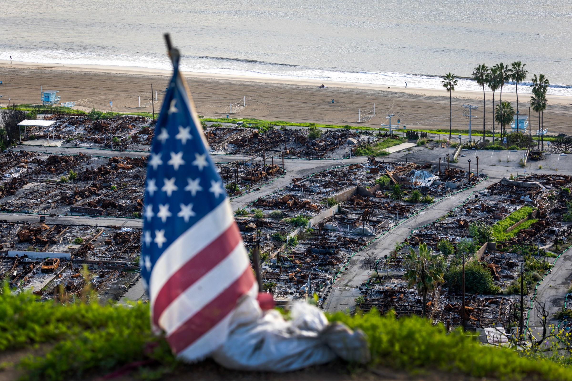 An American flag and sand bag stands on a cliff overlooking the remains of the Pacific Palisades Bowl Mobile Estates, which was destroyed in the 2025 Palisades fire.