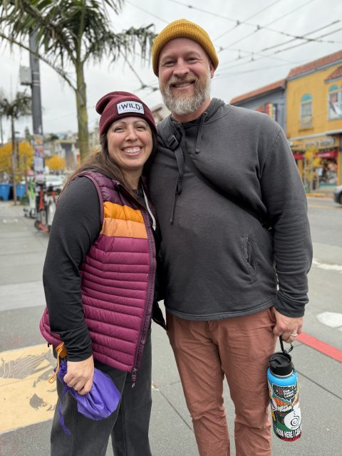 Two people stand side by side on a city sidewalk, smiling at the camera. One holds a water bottle with stickers; both wear hats and casual outdoor clothing. Palm trees and buildings are visible behind them.