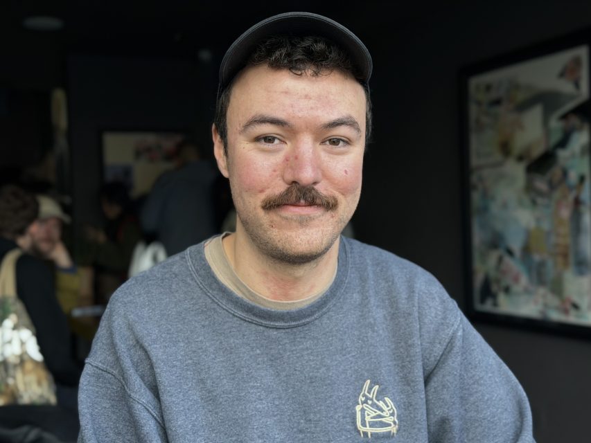 A person with a mustache wearing a gray sweatshirt and black cap sits indoors, looking at the camera. The background is softly blurred.