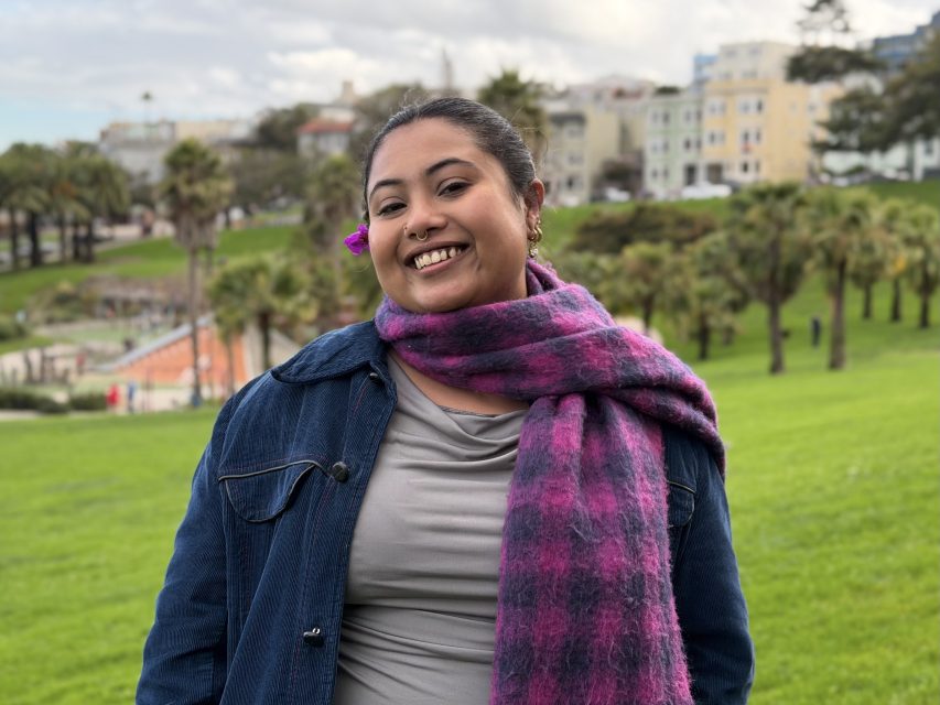 A person smiles outdoors in a park, wearing a denim jacket, gray top, and pink plaid scarf, with a flower in their hair. Buildings and palm trees are visible in the background.