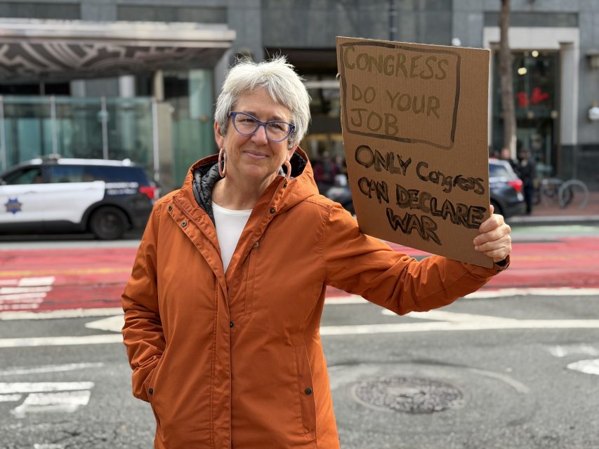 An older woman in an orange jacket holds a cardboard sign reading, "Congress do your job. Only Congress can declare war," standing on a city street near a police car.