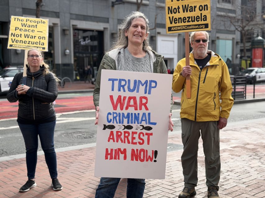 Three people stand at a protest holding signs opposing war in Venezuela; the central sign reads, "TRUMP WAR CRIMINAL ARREST HIM NOW!.
