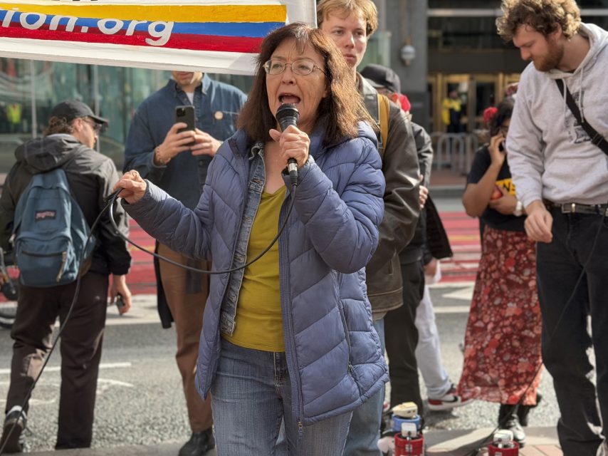 A woman in a blue jacket speaks into a microphone at an outdoor event, with several people standing and listening behind her.