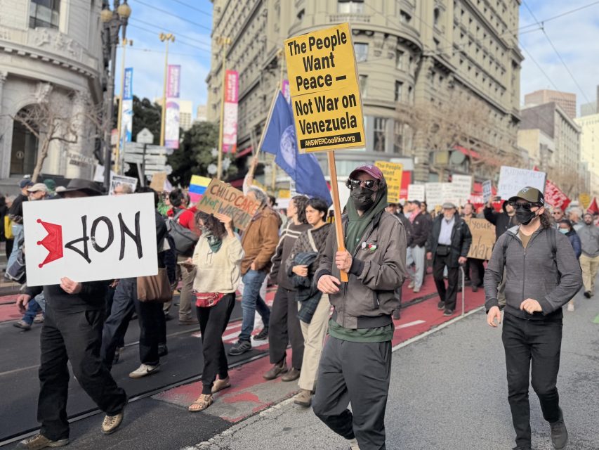 A group of people march down a city street holding protest signs, including one that reads "The People Want Peace — Not War on Venezuela.
