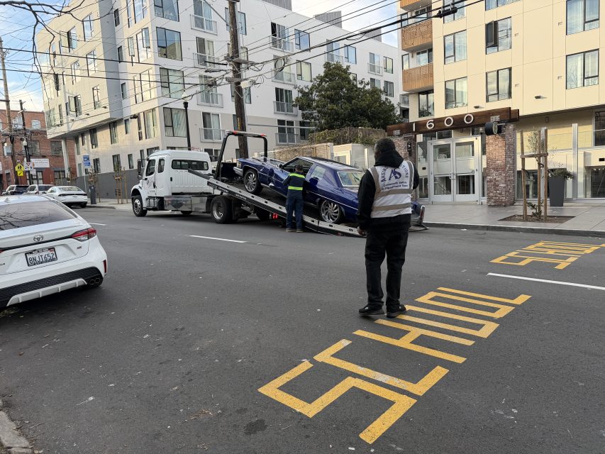 A tow truck loads a damaged car on a city street near apartment buildings, with two workers present and "SCHOOL" painted on the road.