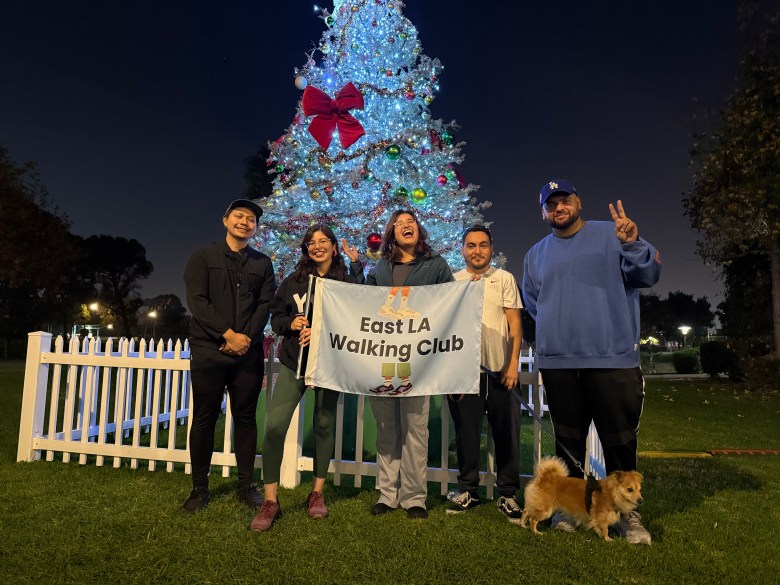 A small group of people hold a flag that reads East LA Walking Club in front of a Christmas tree