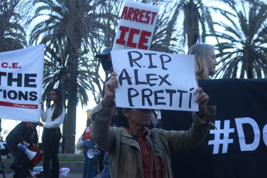 A person holds a sign reading "RIP Alex Pretti" at a protest; other signs in the background say "ARREST ICE" and display hashtags. Palm trees are visible behind the crowd.