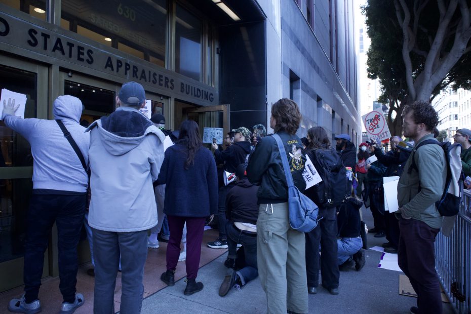 A group of protesters gather outside a building entrance, holding signs and papers, some kneeling and others standing near the doors.