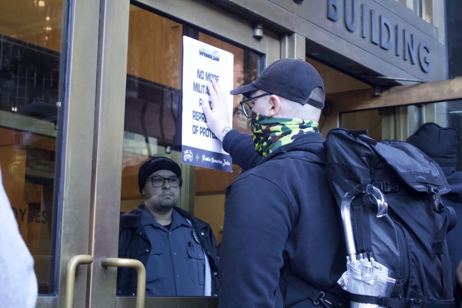 A person tapes a protest sign to a glass door while a security guard watches from inside a building.