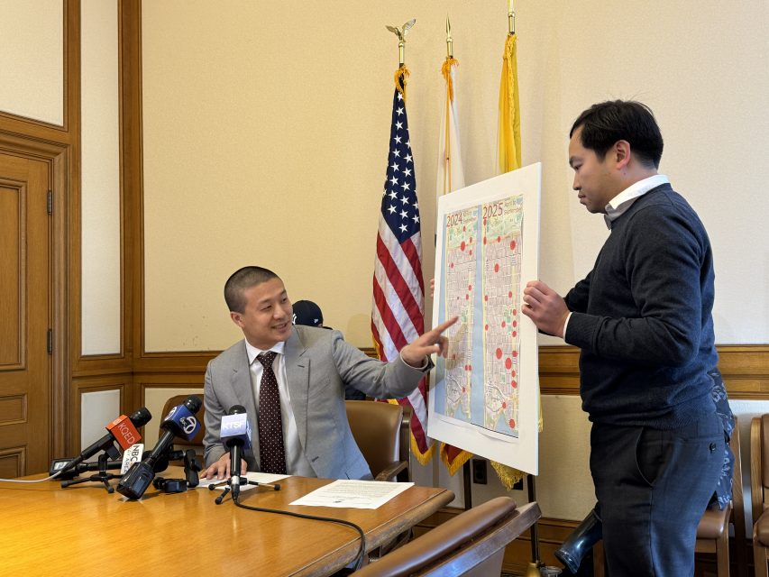 Two men in an office, one seated at a table with microphones, the other standing and holding a large map with colored markings. American and other flags are in the background.