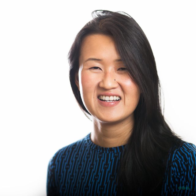 A woman with long dark hair, wearing a blue patterned top, smiles at the camera against a plain white background.