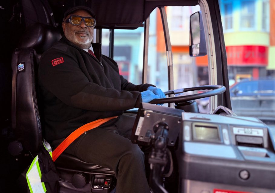 A bus driver wearing a uniform, gloves, and glasses sits at the wheel inside a city bus, with colorful buildings visible through the window.
