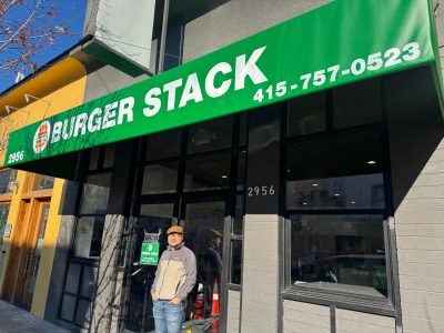 A person stands outside the Burger Stack restaurant at 2956, under a green awning displaying the name and phone number.