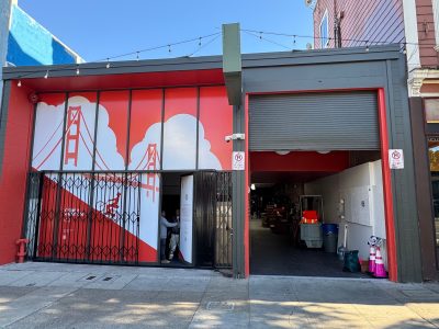 Storefront with a red and white mural of the Golden Gate Bridge, a partially open security gate, and an open garage door revealing items and equipment inside.
