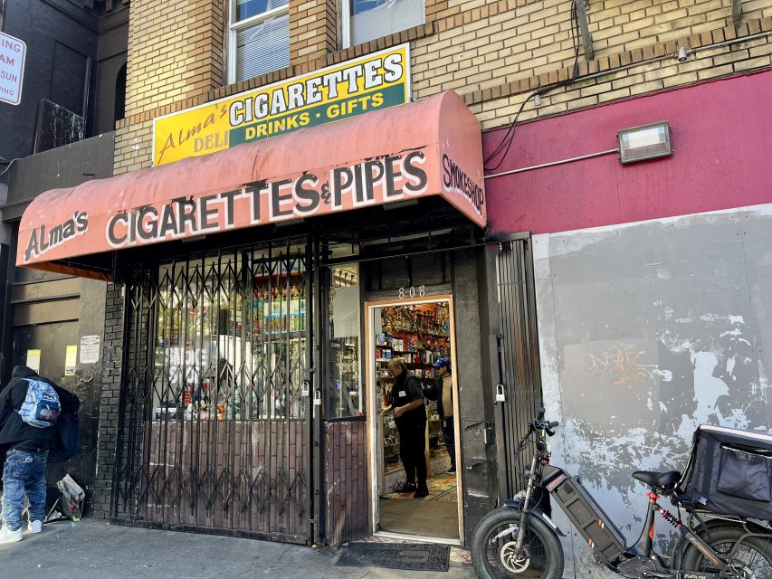 Storefront of "Alma's Cigarettes & Pipes" with a person inside, metal gate partially open, and a bicycle parked outside on a city street.