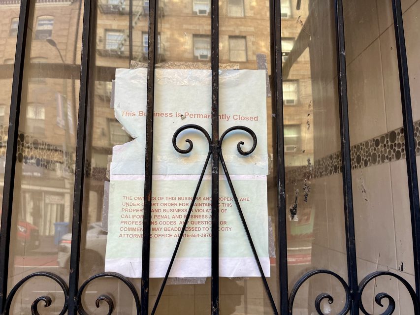 A paper sign taped behind metal bars reads, "This Business is Permanently Closed," with additional text about city attorney contact info and property violations.
