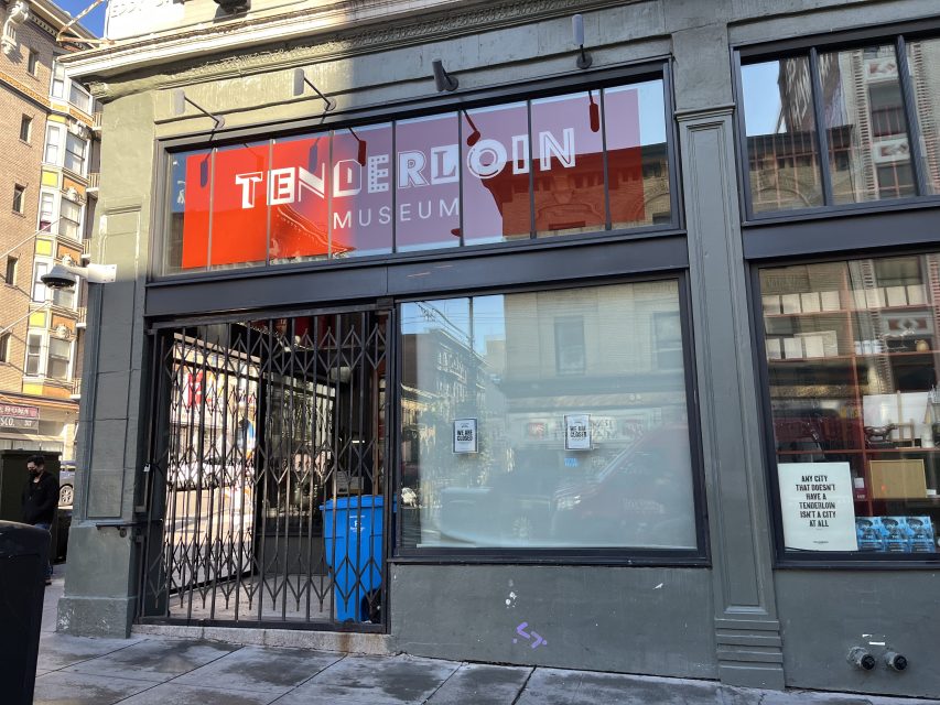 Street view of the Tenderloin Museum with a red sign and gated entrance, located in an urban neighborhood.