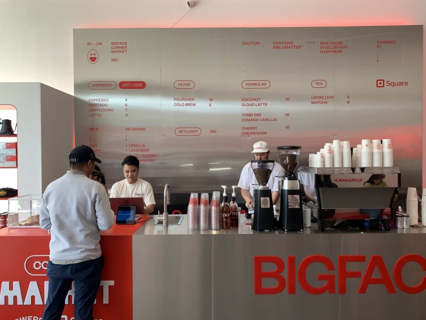 A man orders at a modern coffee counter with two staff members, espresso machines, a menu board, and stacks of cups in a minimalistic café setting.
