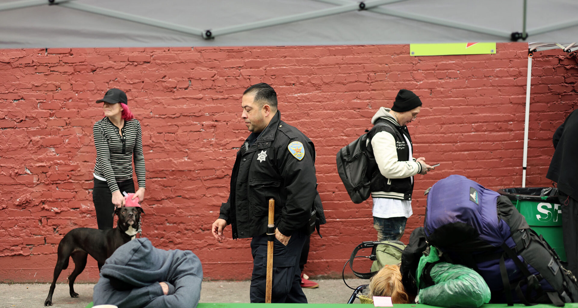 People at an outdoor area: a woman with a dog, an officer walking, a person texting, and another resting on a green table under a canopy. A brick wall serves as the background.