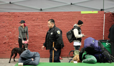 People at an outdoor area: a woman with a dog, an officer walking, a person texting, and another resting on a green table under a canopy. A brick wall serves as the background.