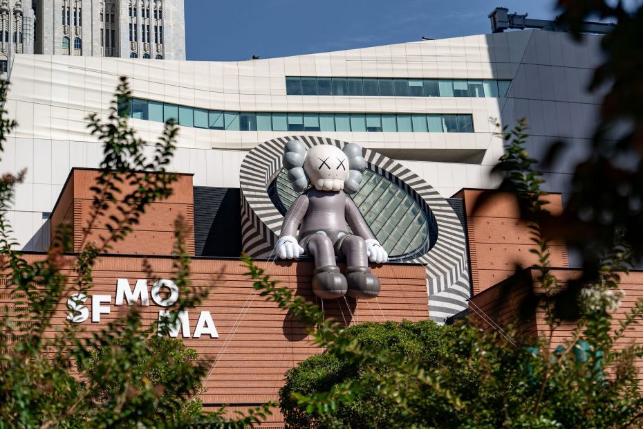 A large gray cartoon-like sculpture with X eyes sits atop the brick facade of SFMOMA, with modern architecture and trees visible in the foreground.