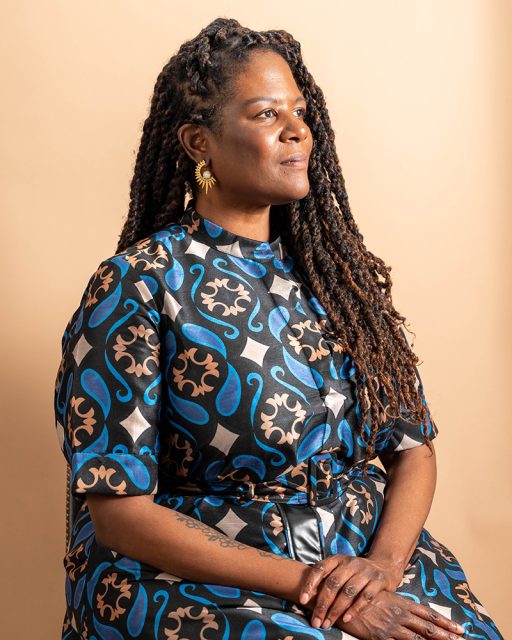 A woman with long, braided hair sits in profile against a beige background, wearing a patterned dress and sun-shaped earrings, with her hands resting on her lap.