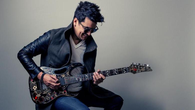 Man wearing sunglasses, black leather jacket, and jeans, seated and playing electric guitar.