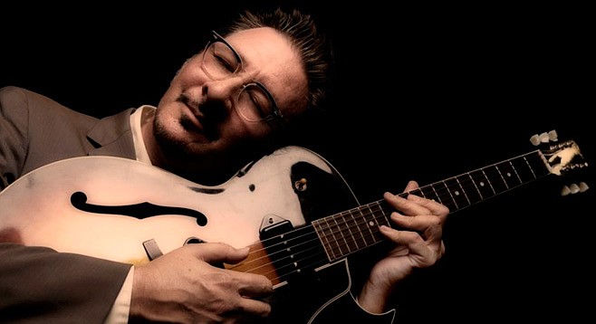 A man in a suit jacket leans his head to the side as he plays electric guitar.