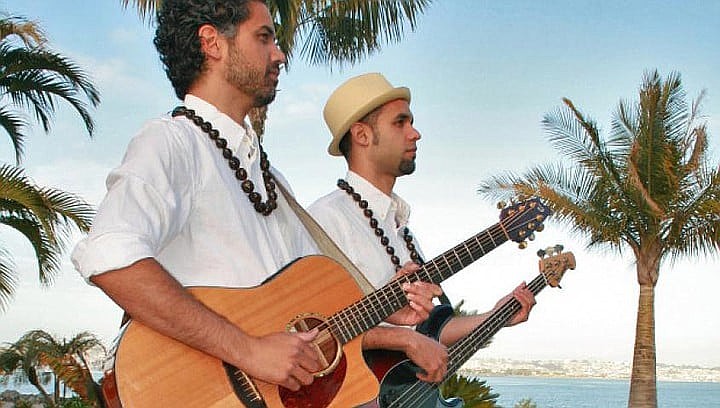 Two men in white dress shirts stand outside by the sea playing guitar