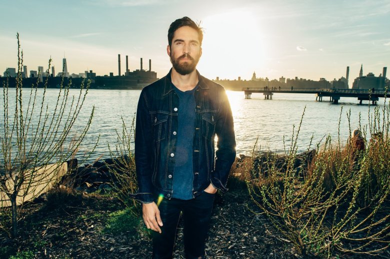 A man with a beard in a jean jacket stands in front of a river at sunset.