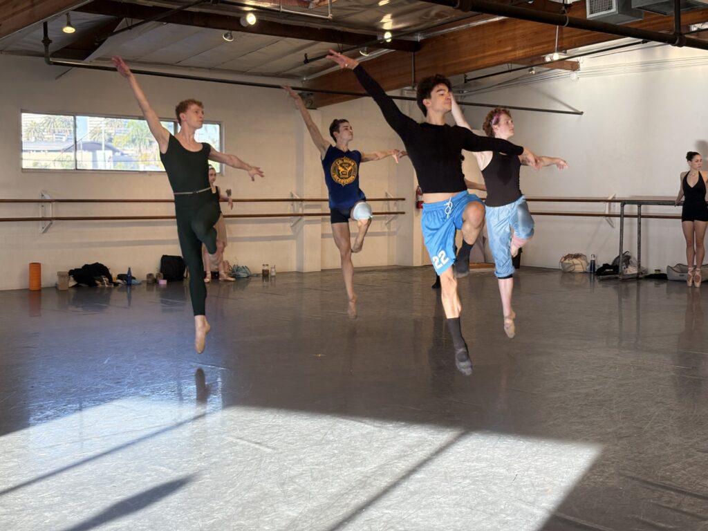 In a ballet studio, four dancers form a box shape and jump in the air with one leg lifted in parallel passé. They lift their arms in an open arabesque line, their torsos turned to face the front of the room.
