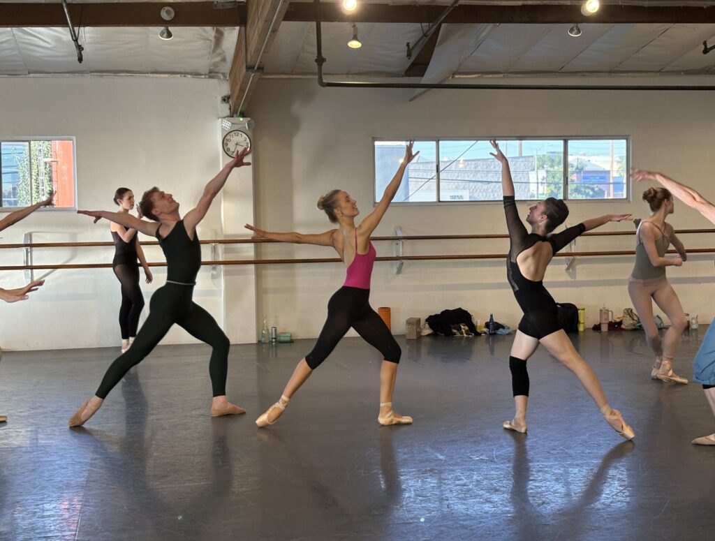 In a ballet studio, dancers rehearse in practice clothes. They form a straight horizontal line, divided in the middle so that the two center women face each other. They pose in tendu derriere with one arm lifted high on the arabesque line.