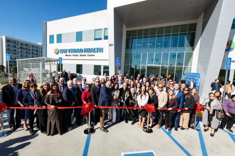 A diverse group of people participates in a ribbon-cutting ceremony in front of the San Ysidro Health: Macias Family Health Center.
