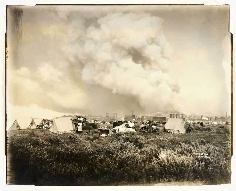 Rows of tents are set up on a grassy field while large clouds of smoke rise from a city in the background, suggesting an emergency or disaster situation.