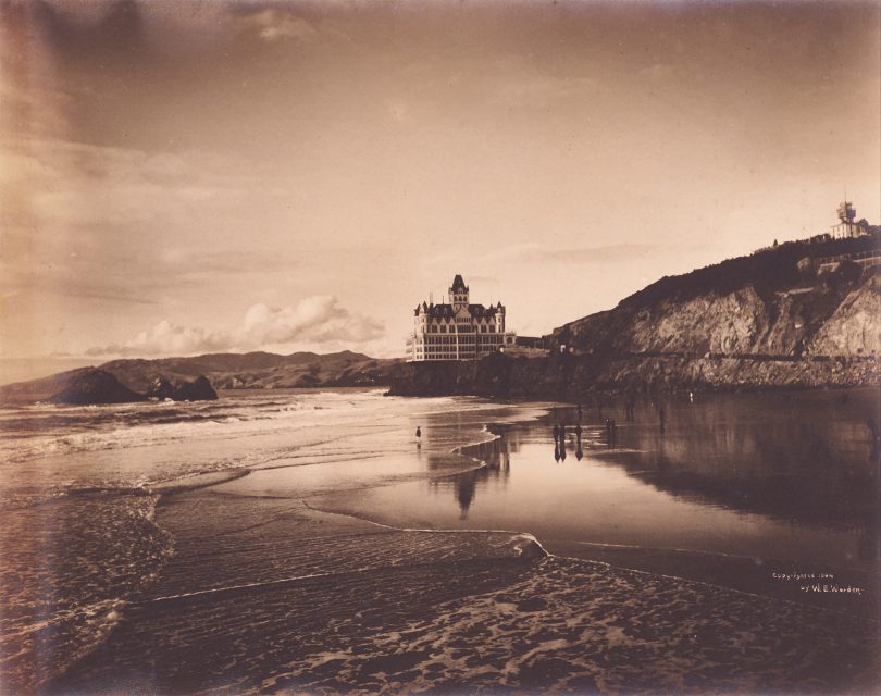 Sepia-toned photo of a large Victorian-style hotel on a cliff overlooking a beach with shallow waves and a few people near the water’s edge.