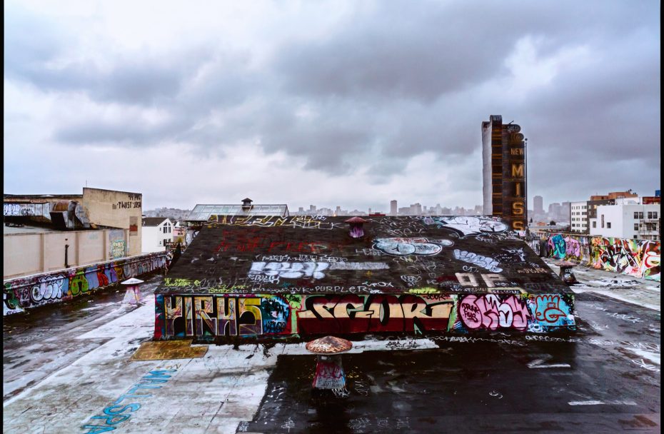Rooftop covered in colorful graffiti under a cloudy sky, with city buildings visible in the background.