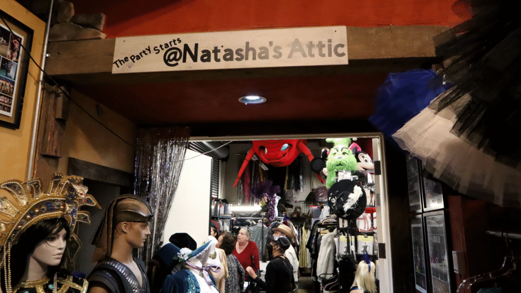 Photo of a sign reading "The party starts at Natasha's Attic" hanging over an open doorway. Parts of costumes are visible around the doorway and in the room, where a handful of people are talking.