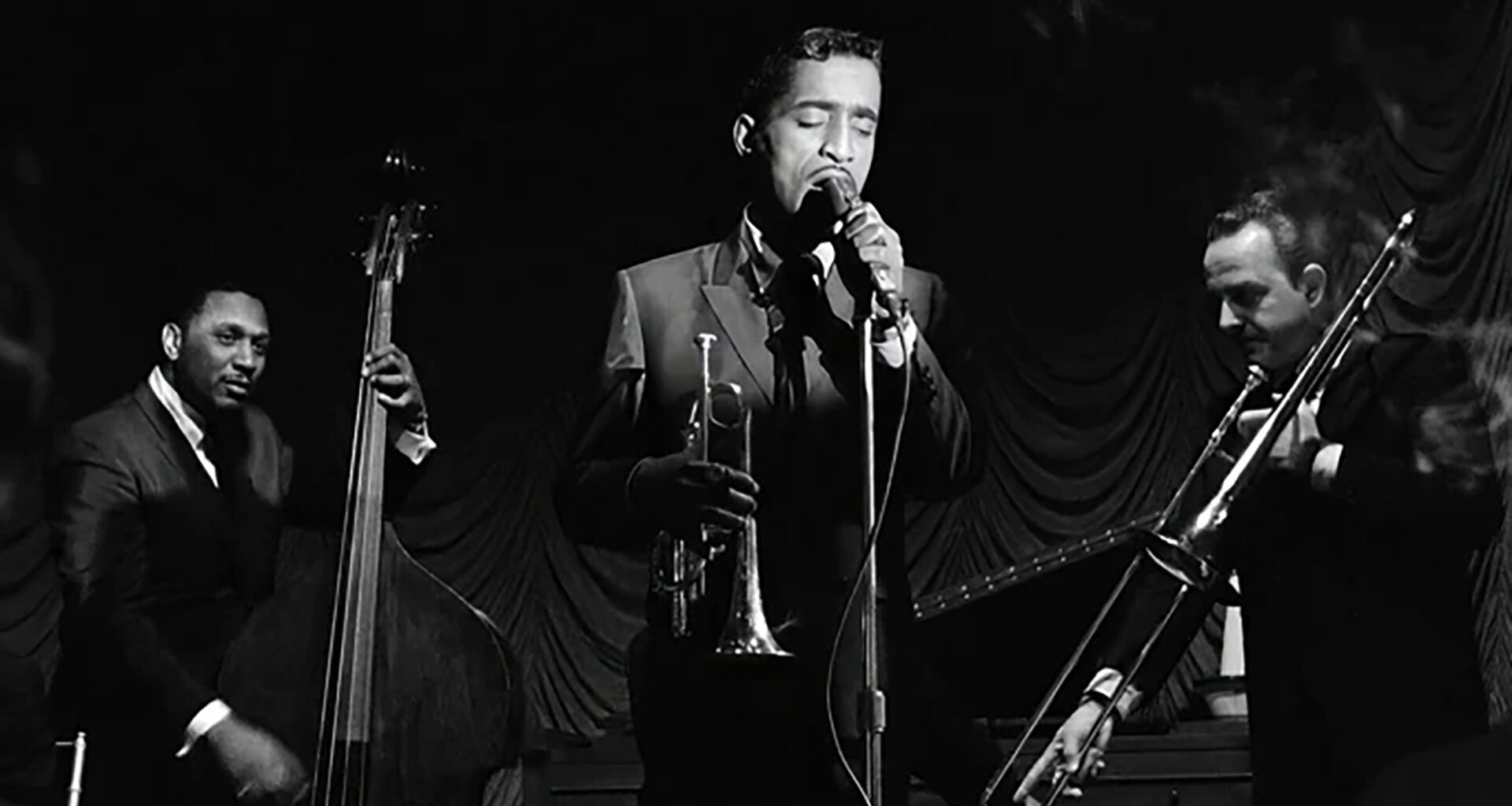 A slendar Black man sings into a microphone, flanked by a bassist and trombonist, while holding a cornet in a black and white image.