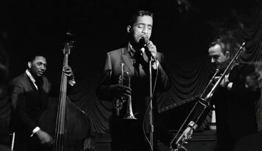 A slendar Black man sings into a microphone, flanked by a bassist and trombonist, while holding a cornet in a black and white image.
