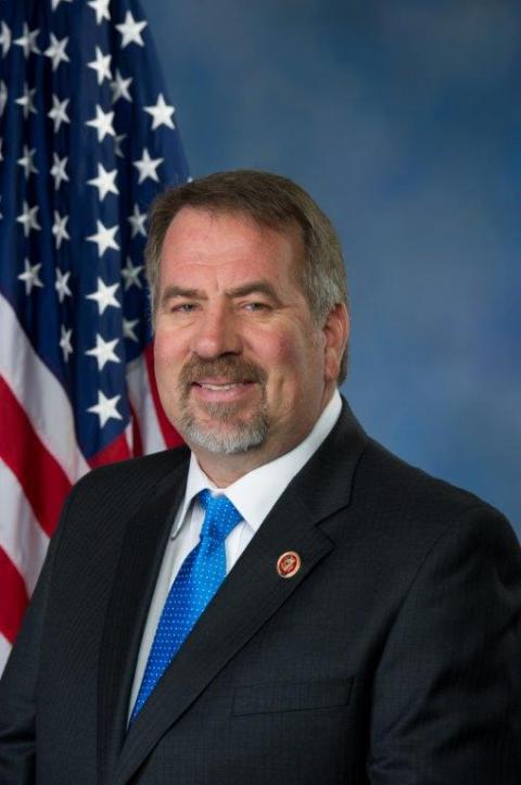 A person with a goatee wearing a dark suit and blue tie, with a U.S. flag in the background
