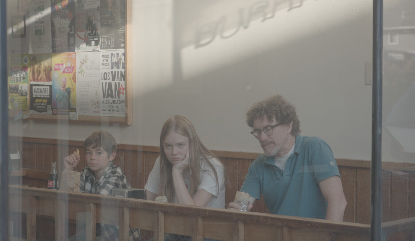 Three people sit at a counter inside a restaurant, each holding food, with neutral or bored expressions. Posters decorate the wall behind them.