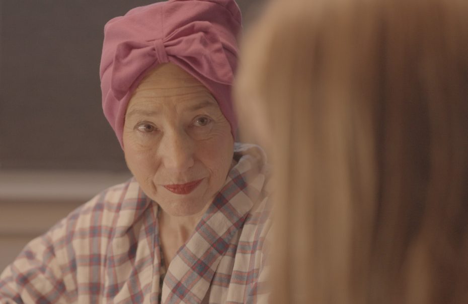 A woman wearing a pink headscarf and plaid robe looks at another person with a slight smile; the scene appears to be indoors.