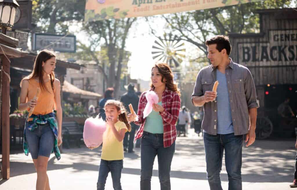 Four people walk together at an outdoor fair, holding snacks such as cotton candy and churros, with shops and trees visible in the background.