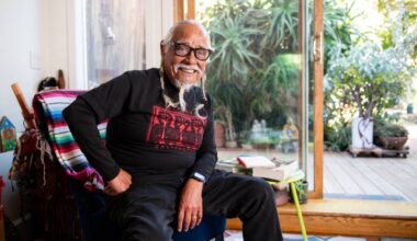 An older man with white braids and glasses sits in a chair smiling, with one arm propped on his hip