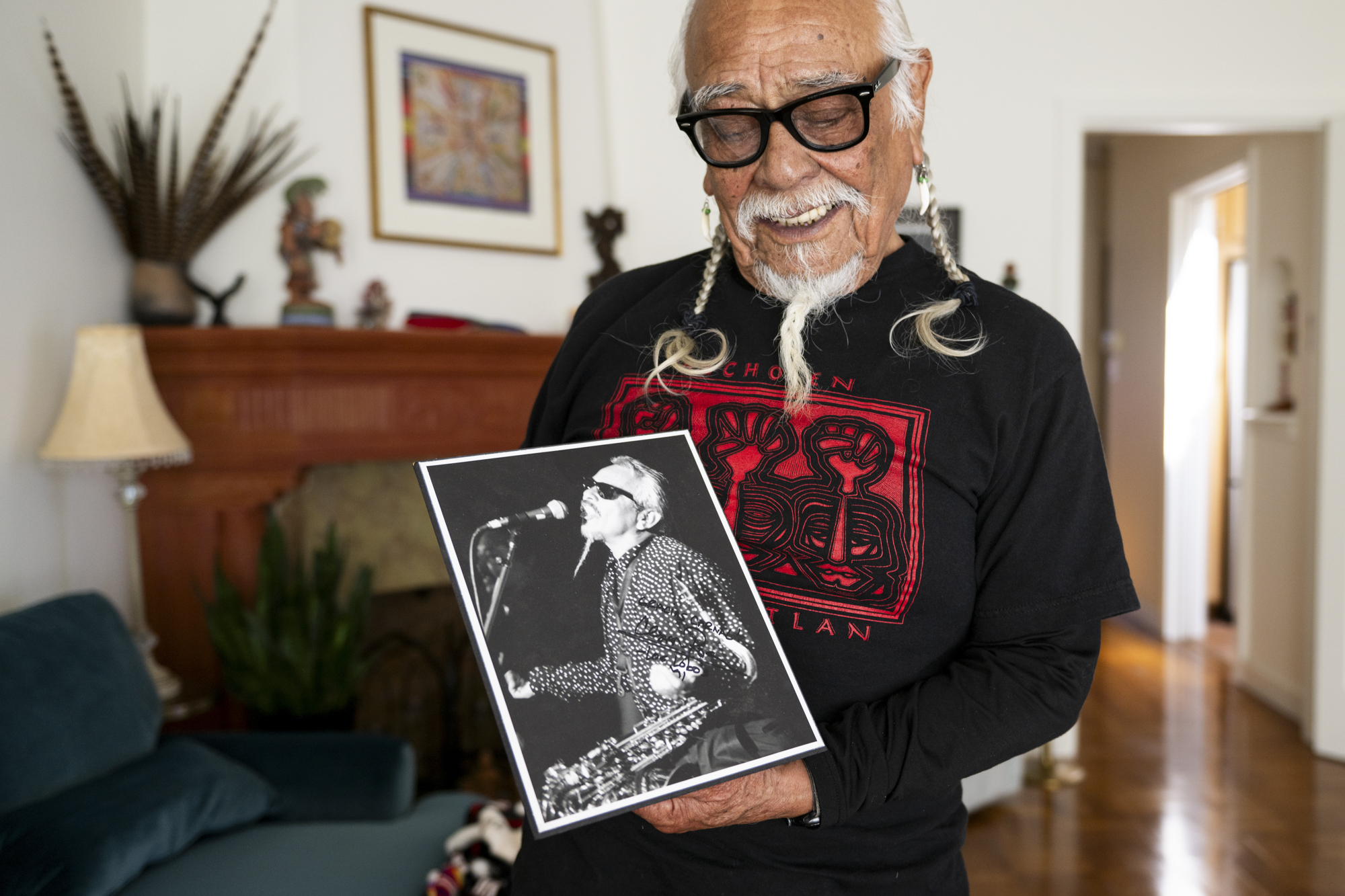 A person with gray hair and a gray moustache and beard holds a photo of himself playing music at an earlier age.