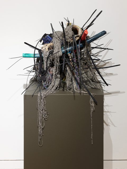 A sculptural assemblage featuring metal rods, fabric, netting, and various found materials arranged on a pedestal against a plain wall.