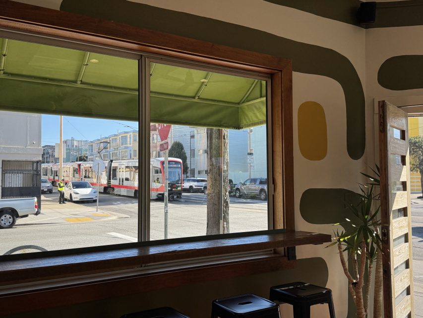 View from inside a cafe looking out a window at a street with passing light rail trains, parked cars, and a person in a yellow vest directing traffic.