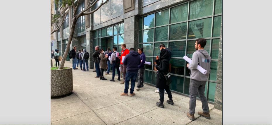 People lined up outside a building, holding documents and blueprints.