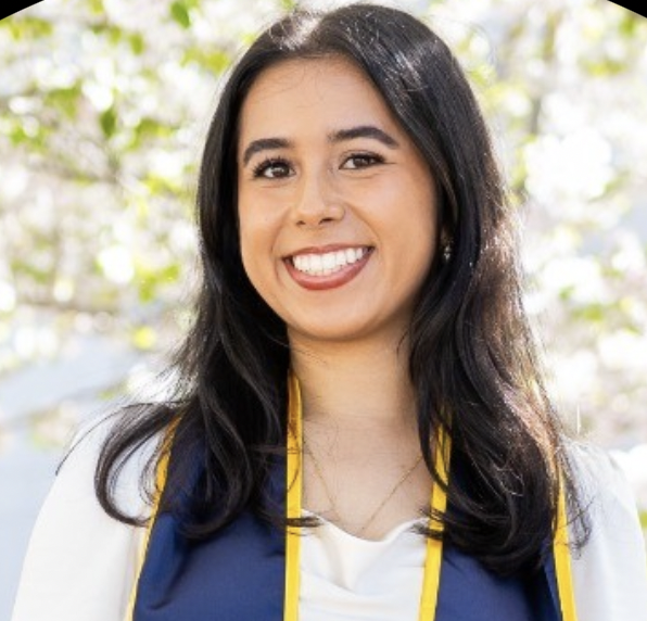 A woman with long dark hair, wearing a white top and blue and yellow graduation stole, smiles in an outdoor setting with blurred greenery in the background.