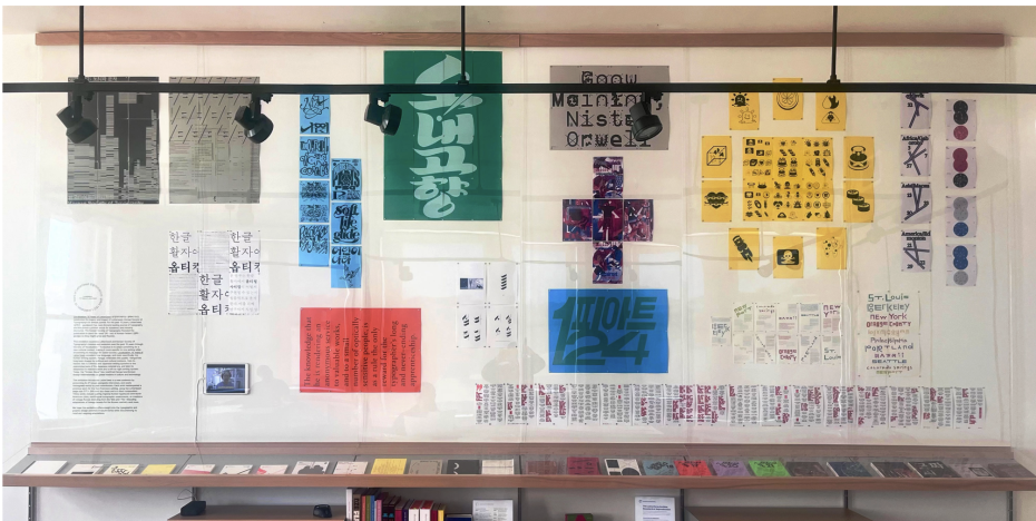 A display case, reminiscent of museums, features colorful posters, zines, and printed papers arranged on a wall and shelf, showcasing graphic designs and typography in various languages.